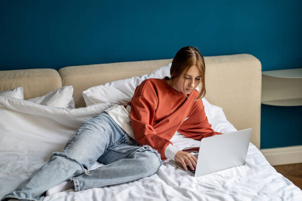 Девочка-подросток, сидящая на ноутбуке и пользующаяся интернетом, отдыхая в спальне. Ребенок-подросток, лежащий в постели с компьютером, болтает с друзьями в социальных сетях, отдыхая дома. Концепция использования технологий для подростков