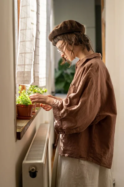 Hipster girl taking care of domestic houseplants, standing by window at home checking leaves and roots of indoors plants. Gardening and plant health care concept.