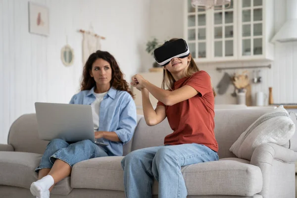 stock image Working from home mother looking at excited little girl daughter wearing VR helmet spending long time immersed in virtual world, selective focus. Dangers of virtual reality for young children