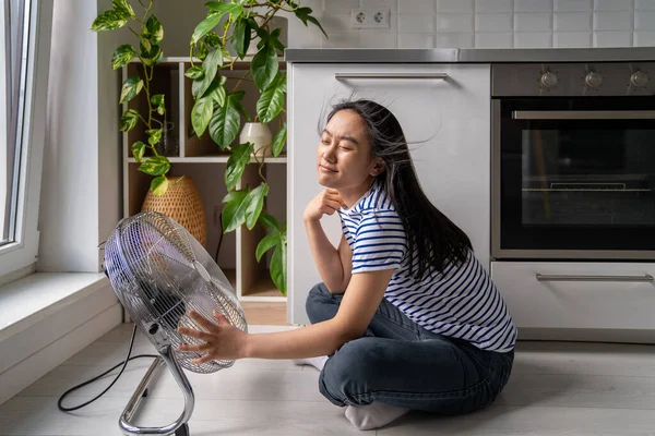 Cheerful Asian girl enjoys cold wind from electric fan sits on floor in kitchen. Young Japanese woman resting at home and sits by fan and enjoys the cool breeze. Summertime, heat concept.