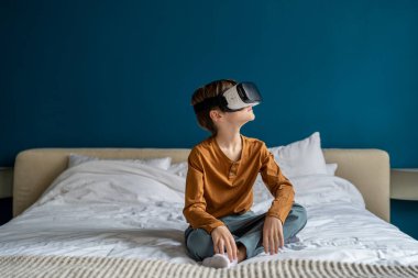 Çocuk sanal gerçeklik kulaklığı kullanarak hayali dünyayla etkileşime giriyor. Beyaz çocuk evde yatağında VR gözlük takmış video oyunları oynuyor. Etkileyici öğrenim ve çocuklar