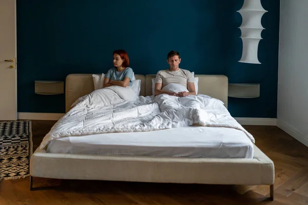 Frustrated sad couple sit on bed under blanket think of relationship problems. Tired offended woman after quarrel lost in thoughts ignores boyfriend with crossed arms. Disappointed lovers in bedroom