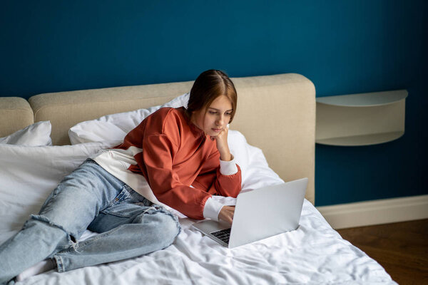 Задумчивая девочка-подросток, лежащая на кровати, использует домашний ноутбук. Заинтересованная школьница набирает текст на компьютере, наслаждается интернетом, дистанционным образованием в колледже, готовится к экзамену на дому