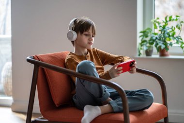 Tranquil kid boy in headphones relax on armchair use phone as video game console. Interested little schoolboy resting watch video podcast on cellphone screen online in headset. Child device addiction clipart