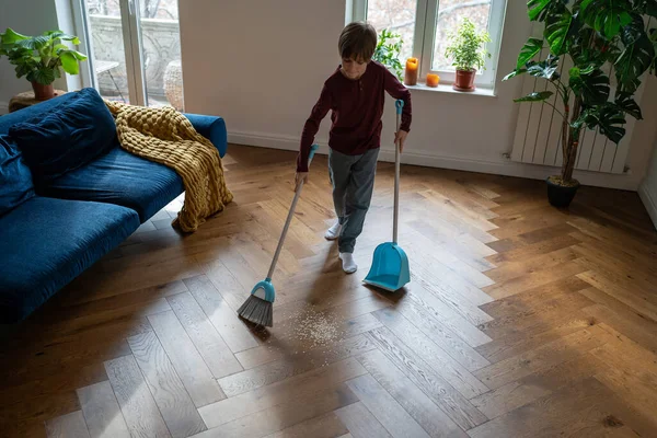 独立した少年 子供の頃からクリーニングハウスの習慣 焦点を当てた少年は ブラシとダストパンでほうきで散乱破片から床を掃引します 子供は家事をする国内清掃 — ストック写真