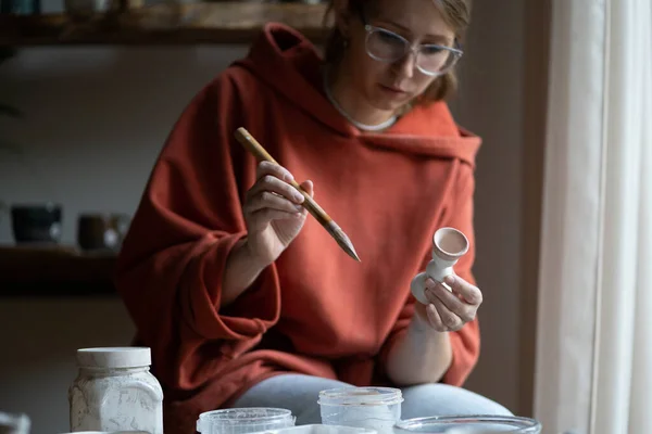 Concentrated creative woman artist enjoys making miniature sculptures from natural clay porcelain. Immersive in process girl sit in ceramic studio with brush at table produces goods for handmade store