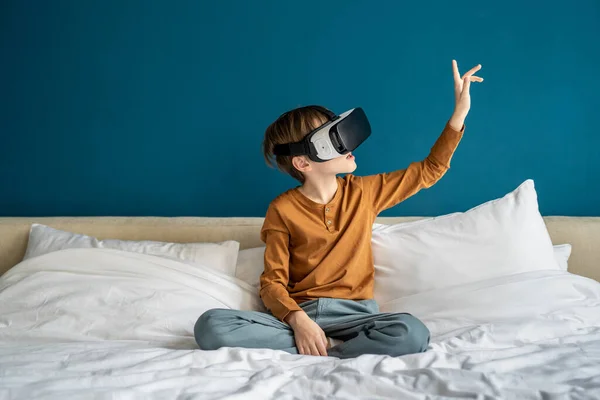 受到启发的男孩正在体验虚拟现实的护目镜 沉浸在未来主义的世界里 有兴趣的小男孩穿着Vr眼镜看智能手机玩电子游戏 用手触摸空气坐在床上 — 图库照片
