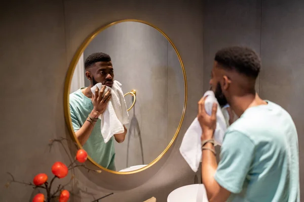 若いアフリカ系アメリカ人の男が洗って顔を拭く不満 男性は化粧品の手順の後にミラーでDermaを調べます 黒男とスキンケアの概念 バスコンセプトの朝のルーチン — ストック写真