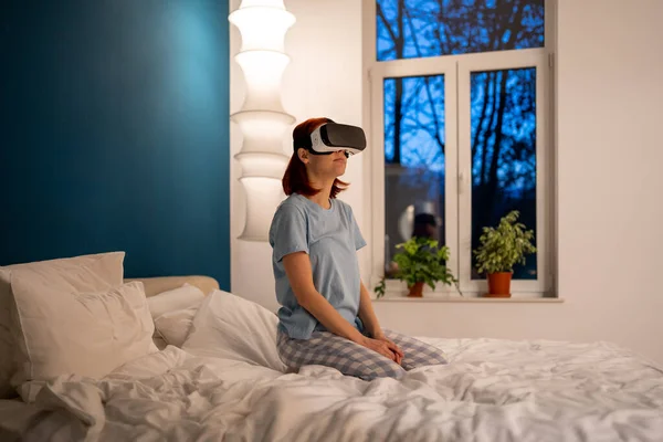 女孩戴着虚拟现实眼镜坐在床上 女人通过特殊的头盔模拟器玩电子游戏 游客穿着特殊的装置在酒店里游览城市 现在的未来 — 图库照片