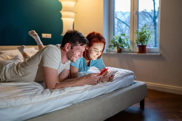 ベッドの中で幸せな夫婦の妻の夫のスマートフォンでは 笑みを浮かべて ベッドの中で横に話している 一緒にオンラインサーフィンソーシャルメディアで面白いビデオを見て 家族の週末家で男の女の夜 — ストック写真