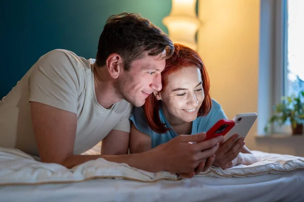 夫婦がベッドに横になって電話を探している 電話で彼氏の面白いビデオを示す女の子 幸せな夫と妻はソーシャルメディアをチェック 男の新しい携帯電話アプリを示す女性 — ストック写真