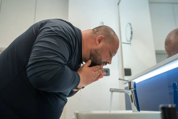 Fat, bald, bearded middle-aged man washes face in bathroom home int sink. Overweight and sweat. Daily routine, hygiene procedure, morning care and body cleanliness concept