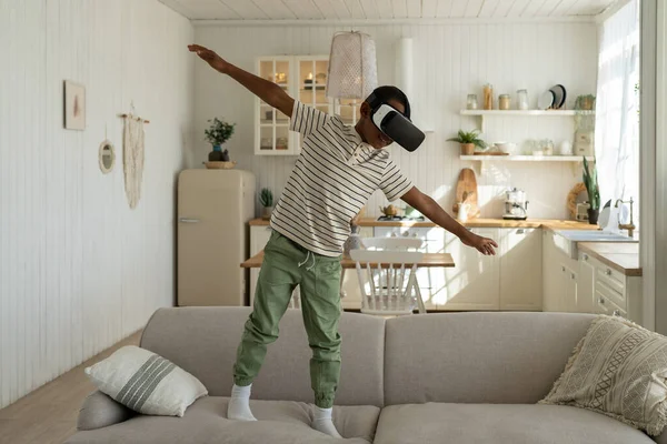 stock image African American boy in 3d virtual reality glasses plays game and fly. Black boy stands on couch and waving hands in helmet simulator. Schoolboy is study planes with help of virtual reality glasses