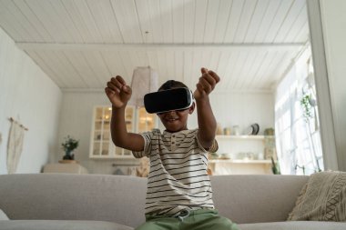 Üç boyutlu sanal gerçeklik gözlüğü takan küçük Afrikalı Amerikalı çocuk evdeki kanepede oturuyor. Çevrimiçi dünya simülasyonu ve oyun, yarış arabası. VR gözlüklü, hava gözlüklü, el kol hareketi yapan ergen.