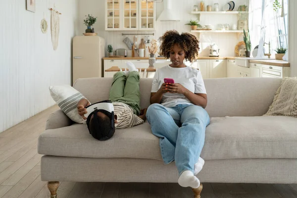 アフリカ系アメリカ人 母親と息子はデジタルデバイスでソファの上で家をリラックスし 母親は仮想現実ヘルメットのゲームをプレイ中の男の子と 携帯電話を使用しています 子供中毒装置 — ストック写真