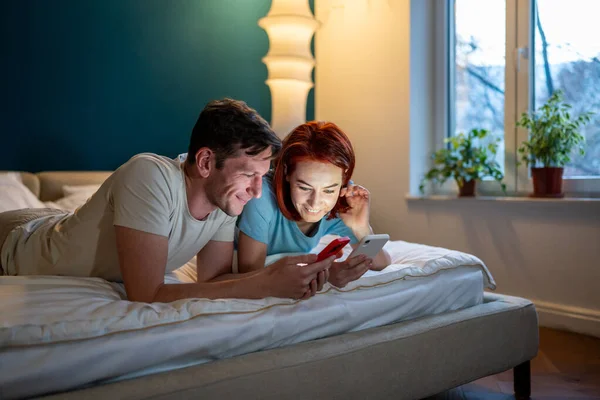 夫婦がベッドに横になって電話を探している 幸せなボーイフレンドとガールフレンドは 携帯電話や笑顔で面白いビデオを見て ソーシャルメディアをチェックします 女性の新しい携帯電話アプリを示す男 — ストック写真
