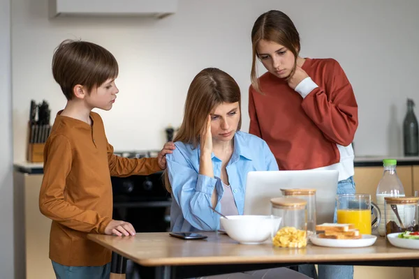 头痛的妈妈在厨房里工作笔记本电脑 孩子们站在屏幕旁边看 厌倦了 精疲力竭的工作妇女在家里做着遥远的工作 抚养着儿子的女儿 母亲和职业概念 — 图库照片