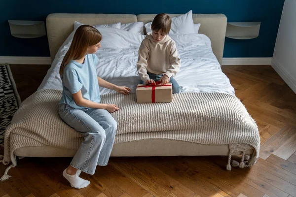 小さな男の子の子供が開いたベッドの上に座っている母親は 自宅で一緒に誕生日を祝うプレゼントボックス お母さんと小さな子供を包みました 予期せぬ贈り物で親の驚くべき息子を愛する — ストック写真