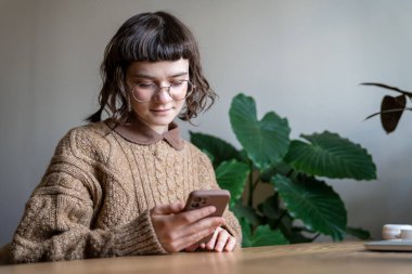 Gözlüklü genç kız evde dinlenirken akıllı telefon kullanıyor, masada oturmuş mesaj mesajı okuyor. Sosyal medya paylaşımlarında gezinen, arkadaşlarıyla sohbet eden, telefon ekranına bakan genç