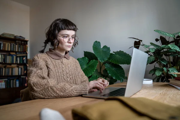 ノートパソコンでテーブルに座っている眼鏡をかけ 自宅からオンラインで勉強しながらコンピュータ上の情報を検索する10代の学生の女の子に焦点を当てました 距離学習 技術とティーンエイジャー — ストック写真