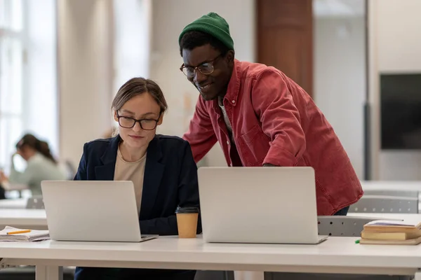 Smiling african american guy helps caucasian girl students working on project in college university library. Positive man woman look at laptop screen explaining studying learning together in campus.