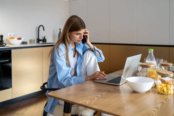 忙碌的女自由撰稿人在家里厨房的笔记本电脑上有智能手机对话 遥远的工作理念 年轻的高加索女商人在电脑电话上工作 — 图库照片