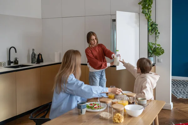 stock image Happy family breakfast together in kitchen. Caring teenage girl hands small brother bottle of milk from refrigerator for cornflakes. Loving mother help to son with healthy food at home. Enjoy morning