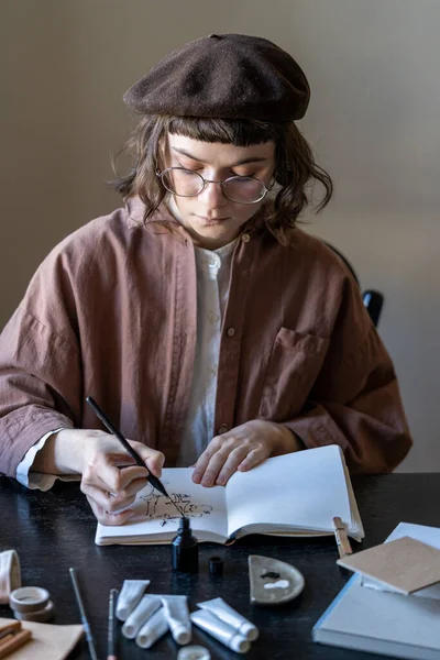 Mladá Zaostřená Umělkyně Baretu Brýlích Sedí Stolní Malby Černým Inkoustem — Stock fotografie