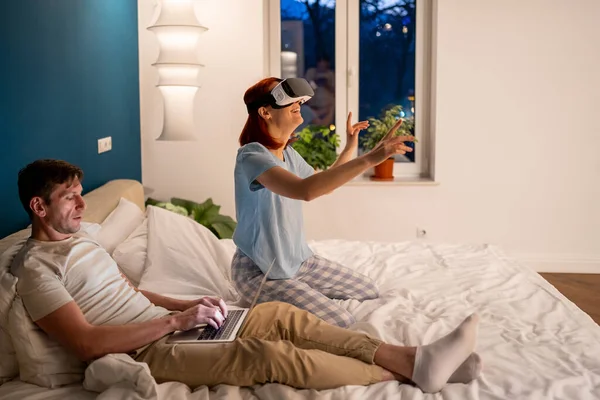 专注的男朋友坐在床上 带着Vr 3D眼镜和女友一起玩电子游戏 感兴趣的男人通过计算机控制妻子的虚拟现实头盔 今后在国内使用的技术 — 图库照片