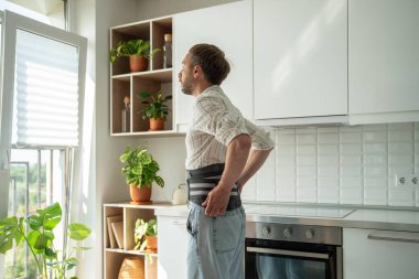 Man in sick leave at home on kitchen wearing back support belt corset on lower back during exacerbation to treatment of hernia, postoperative recovery. Back pain, spine health problems concept.  clipart