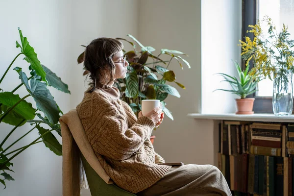 富有想像力的梦想女孩坐在扶手椅上 手中拿着一杯咖啡 望着窗外 在舒适的房子里享受着一天的时光 把绿色的植物放在罐子里 年轻女人想到了生活 精神健康概念 — 图库照片