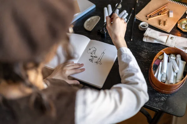 Illustrator hands drawing in notebook with black ink fantasy art. Designer freelancer preparing order in studio. Creative hobby, profession. Aesthetic workplace for inspiration for new project.