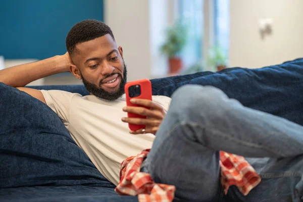 リラックスしたアフリカ系アメリカ人の笑顔の男がソファに座って リビングルームの自宅でスマートフォンを見ています 面白いビデオや冗談で笑ったり ソーシャルネットワークで友達とチャットしたり — ストック写真