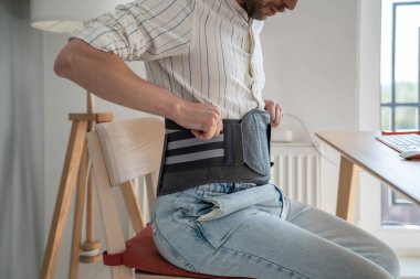 Man sitting on chair wearing back brace to reduce hernias, using decompression corset for herniated discs at home, relieving symptoms of degenerative disc disorder. Recovery after lower back surgery clipart