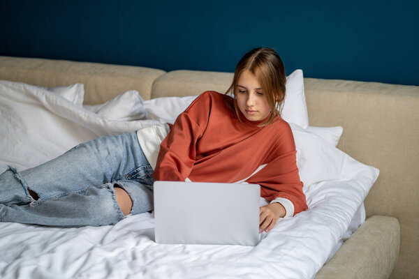 Задумчивая девочка лежит расслабленной на кровати с ноутбуком, печатающим на клавиатуре, общается в социальных сетях выбирает колледж для поступления. Интернет-зависимость среди подростков, отсутствие интереса у студентов