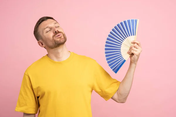 stock image Overheated man using paper fan suffer from heat, sweating, cools herself due problem no air conditioner at home at summer weather, enjoying fresh air and closed eyes isolated on studio pink background