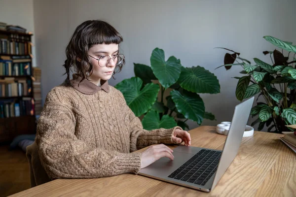 ノートパソコンでテーブルに座っている眼鏡をかけ 自宅からオンラインで勉強しながらコンピュータ上の情報を検索する10代の学生の女の子に焦点を当てました 距離学習 技術とティーンエイジャー — ストック写真