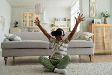 VR kulaklık takan neşeli küçük Afrikalı Amerikalı çocuk boş zamanlarını sanal gerçeklikte geçiriyor. Mutlu çocuk oturma odasında oturmuş online oyun simülasyonu oynuyor, AR 'ı keşfediyor.