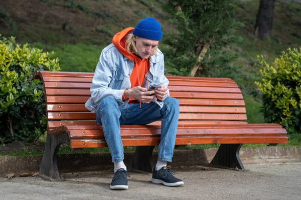 公園のベンチに一人で座って スマートフォンを持って 悪いニュースを読んでいる若い男 良いテキストメッセージを受け取らず 電話で女の子と別れ 悲しみと心の傷を感じる — ストック写真