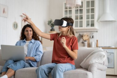 Annesi uzaktan kumandalı bilgisayarla çalışırken kızı da VR gözlüğü takıyor oturma odasında kanepede yan yana oturuyor. Sanal gerçeklik kulaklığıyla oynayan bir kız. Gelecekteki teknolojiler ve çocuklar