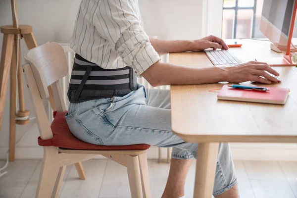 stock image Freelancer man works types on computer keyboard wears back support belt corset on lower back to relieve stress on spine. Treatment of hernia. Back pain health problems, consequences sedentary work. 