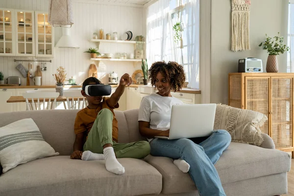 African American family mom and son sit on sofa with electronic devices. Mother-parent sit with laptop, son in virtual reality glasses. Black boy show with hand in gaming helmet. Family and technology