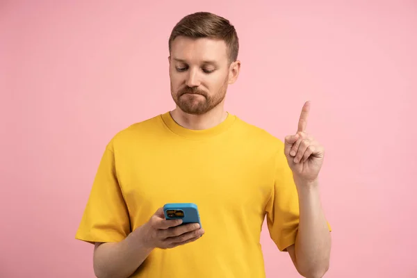 悲しいことに 男性は指数を指摘し ピンクの背景にあるスマートフォン画面に表示されます 黄色いTシャツのひげ付き男性は 携帯電話の読書情報に不満を抱いていました 悪いニュース トラブル — ストック写真