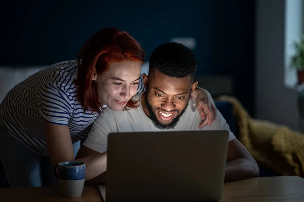 Happy married multiracial couple using laptop together, planning summer vacation or shopping online. White woman and African American man choosing movie to watch in evening at home, make a video call