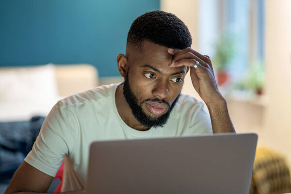 Focused African American guy freelancer using laptop at home, overworked black man working on project all night at home office, student guy remote studying looks at computer screen feeling tired