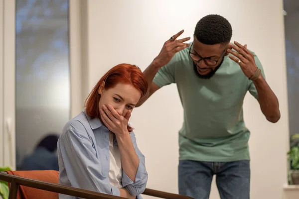 stock image Mad angry African American man husband blaming shaming wife, young woman sitting on chair at home and crying while being being emotionally abused by partner. Multiracial couple having fight at home