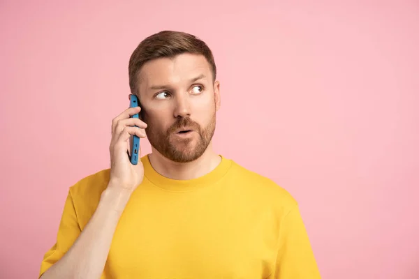 スタジオのピンクの背景で電話で話している黄色いTシャツのイライラした男を困惑させた 驚いた男性が外を見上げてスマートフォンを呼ぶ ハードコミュニケーション 会話の問題 悪いニュース — ストック写真