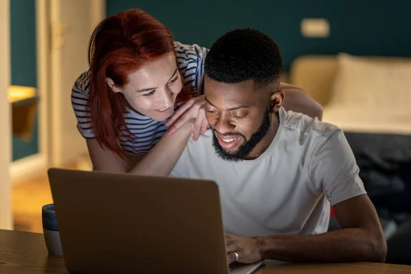 Loving young caucasian woman talking with boyfriend working on laptop late at home, smiling girlfriend demanding attention from busy with work African American guy freelancer. Work life balance