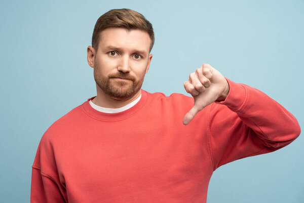 Displeased irritated sad man showing thumb down dislike gesture isolated on blue background looking at camera with regret, studio portrait. Negative assessment, review. Sincere human emotions concept.