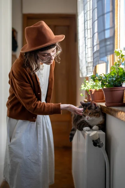 Caring girl gently touches fluffy cat lying on radiator under windowsill with houseplants in pots at sunny home. Pet lovers. Affectionate kitten lies on radiator by window with young woman mistress